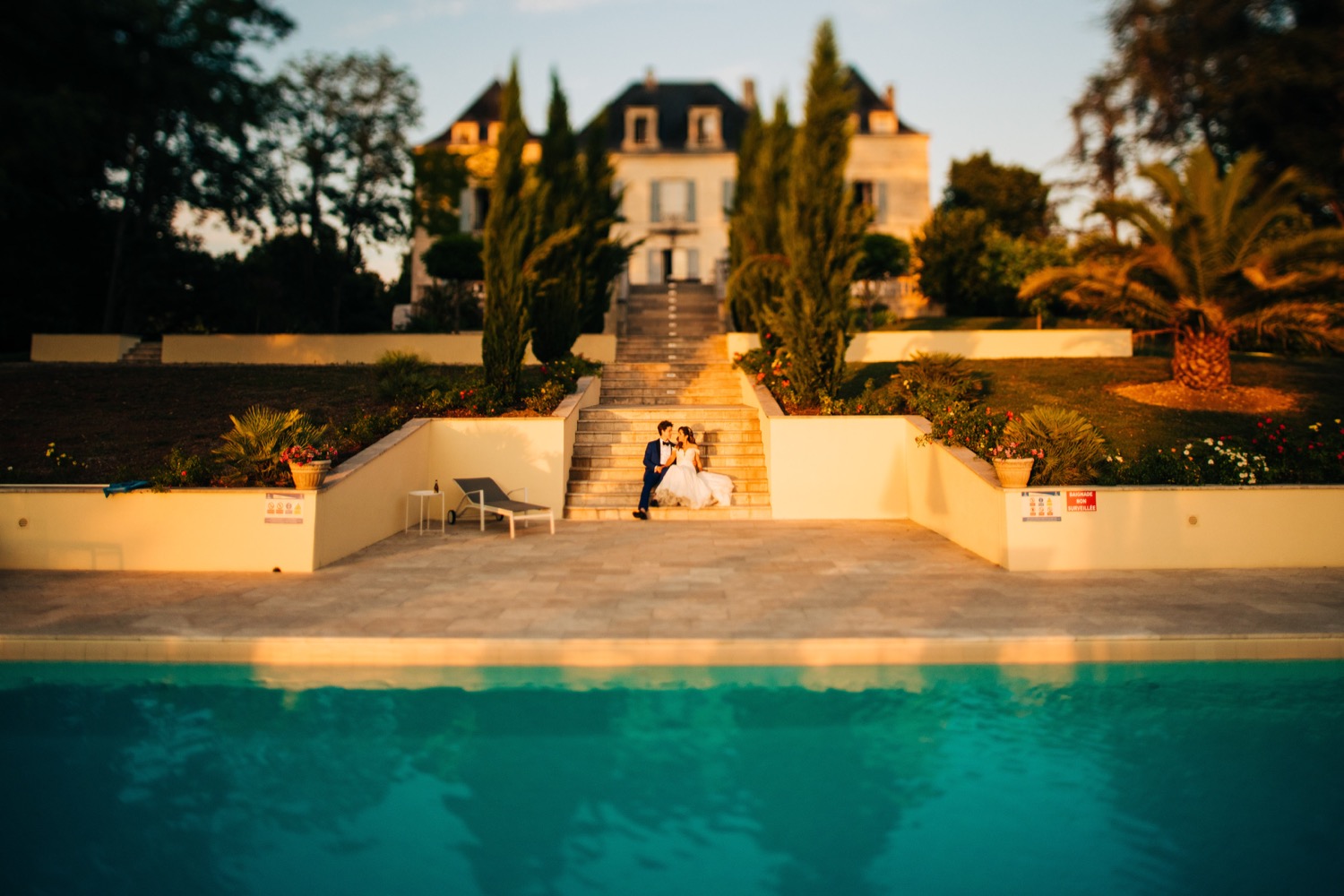 A bride and groom sit on the stairs between the outdoor pool and mansion at Domaine La Fauconnie, a wedding venue in France, as Magic Wedding Photographer takes their photo.